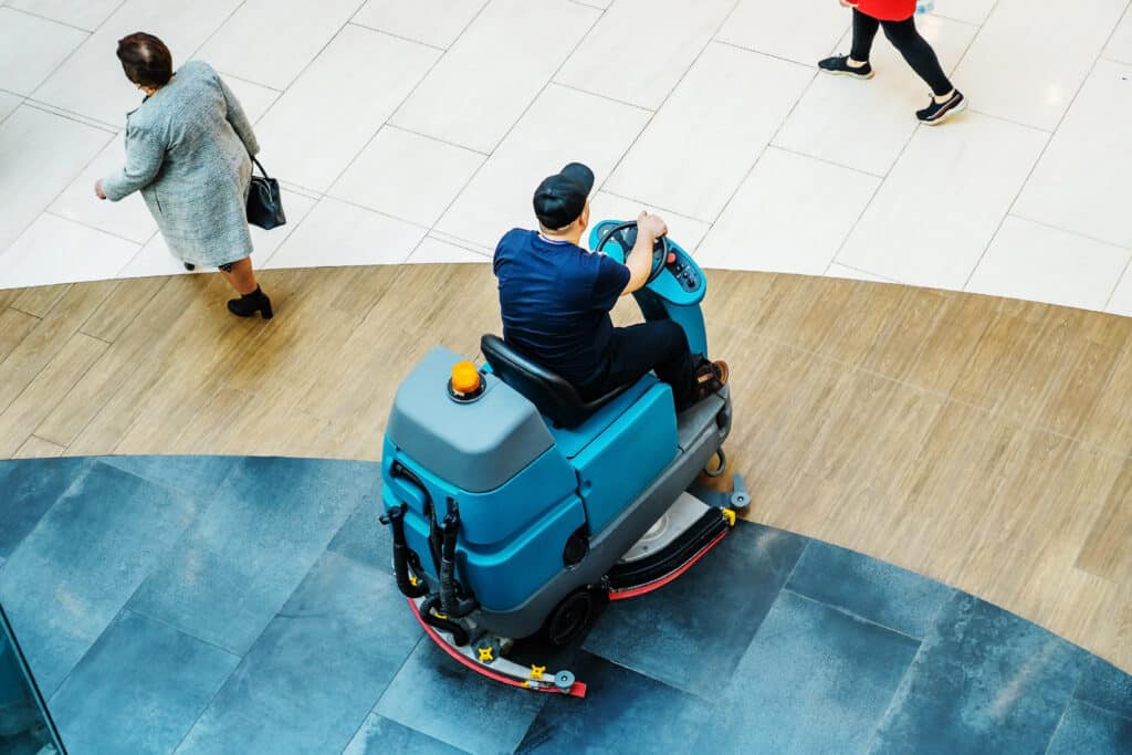Autolaveuse bleue professionnelle nettoyant un sol carrelé dans un centre commercial. Un agent et des passants sont visibles.