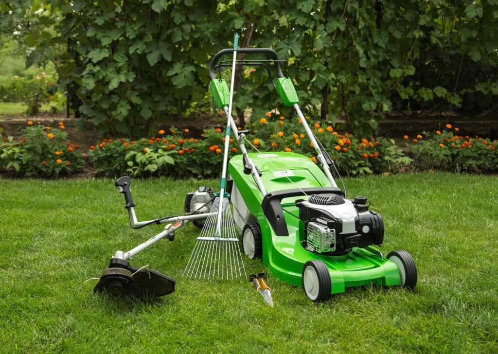 Tondeuse à gazon verte, débroussailleuse, rateau et outils sur pelouse verte, fond de buissons et fleurs oranges.
